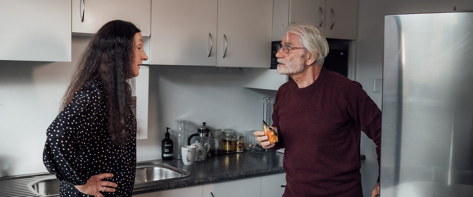 An older man getting angry with a woman with long dark hair, standing in a kitchen.