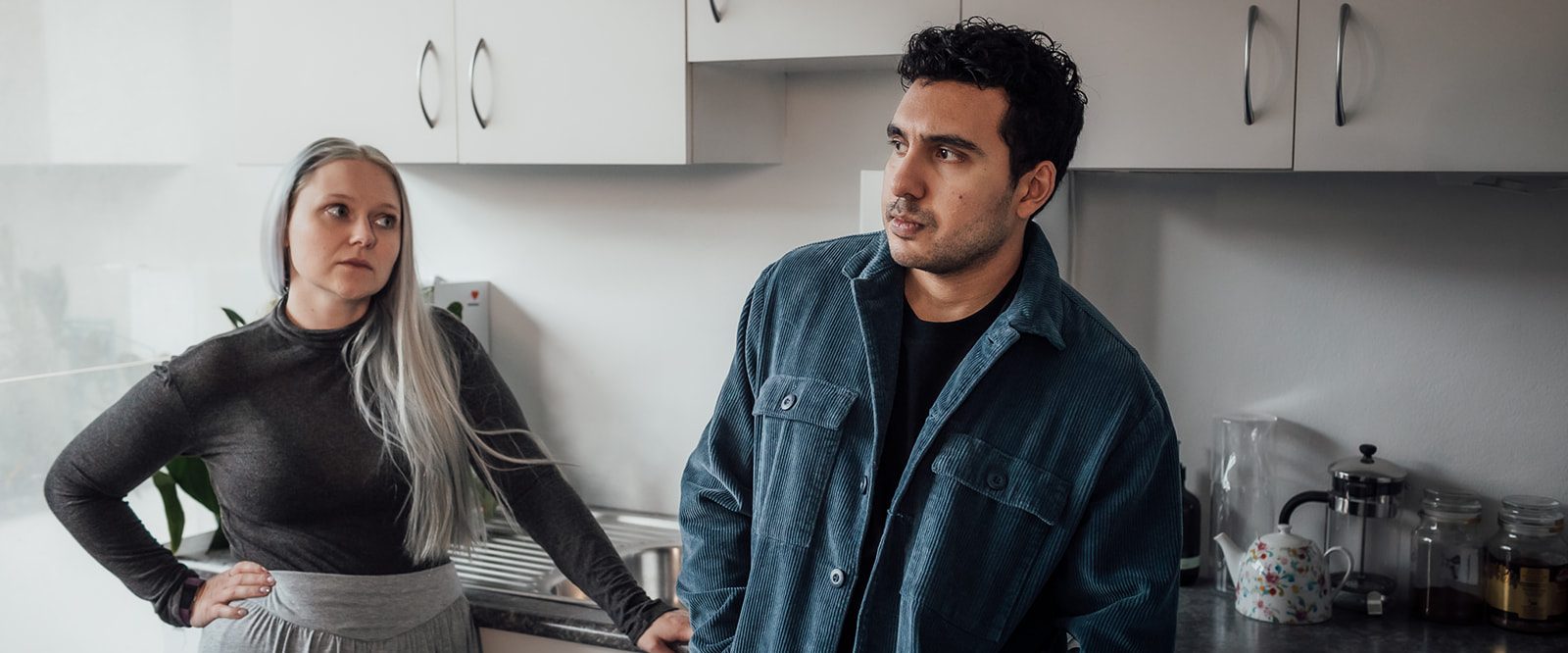 A woman and man standing in a kitchen, looking annoyed with each other.