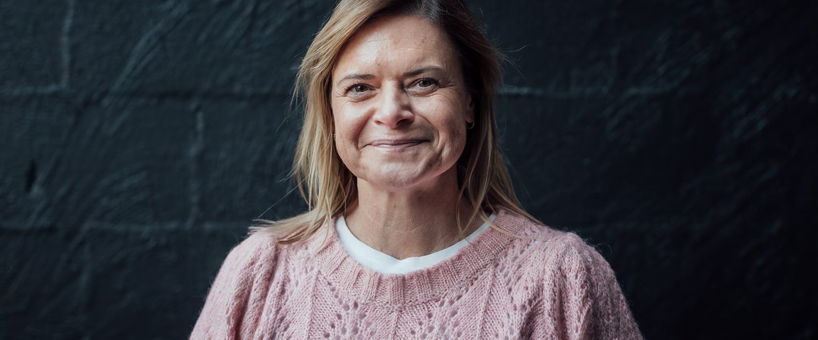 A smiling woman in a pink woolen jumper standing in front of a black brick wall.