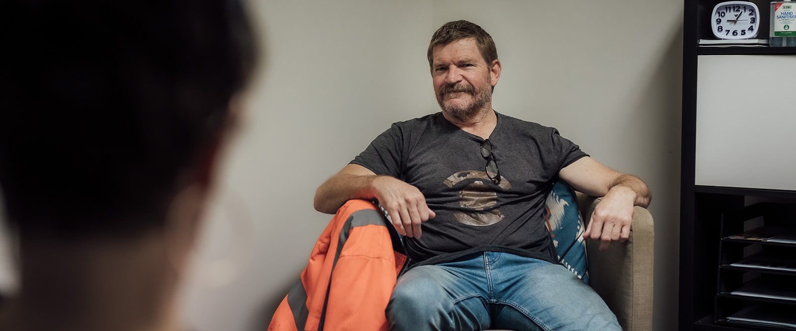 An unshaven man sitting in an armchair in front of a coffee table, talking with a person.