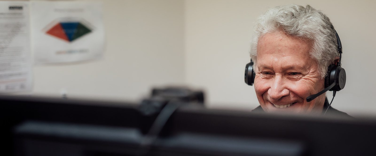A smiling man sitting at a computer, talking on a phone headset.