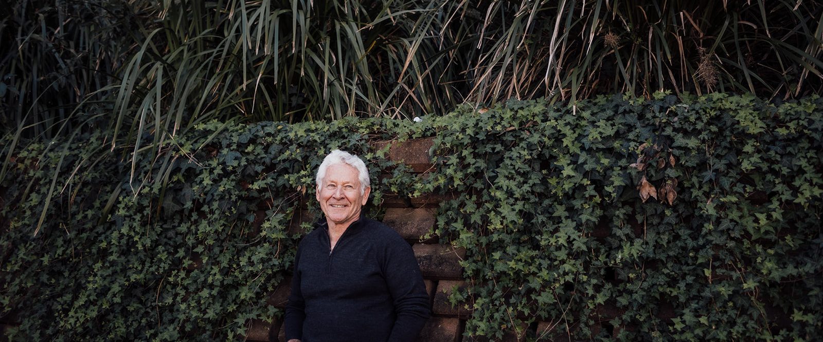 An older man smiling thoughtfully, leaning on a brick wall covered in ivy.