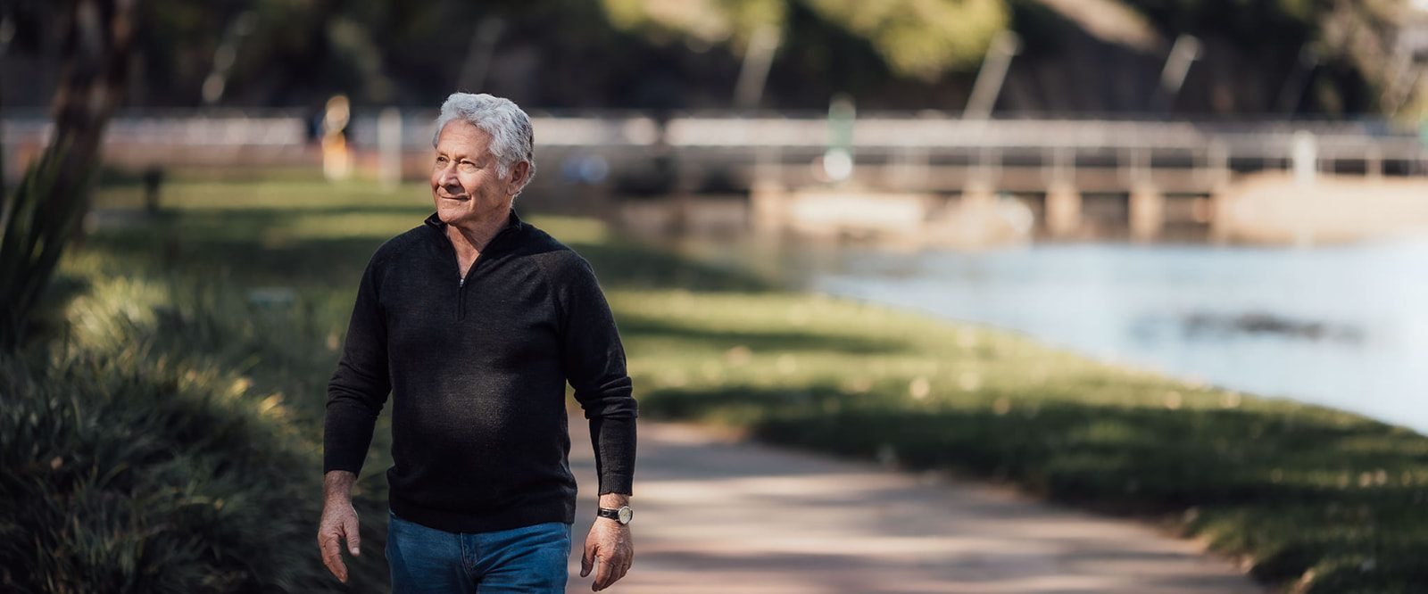 A smiling older man walking on a path by a river.