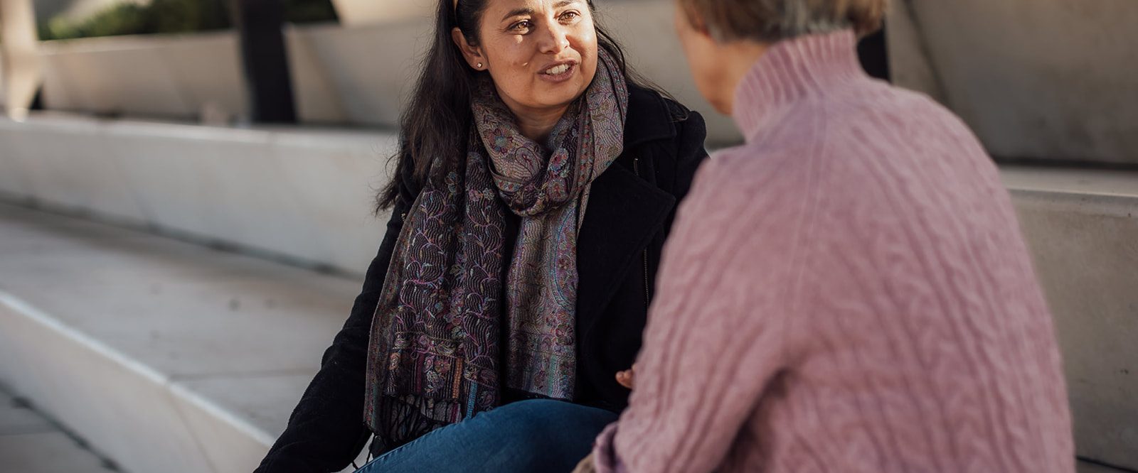 Two relaxed women sitting with the sun on their faces talking together on the steps in a park.