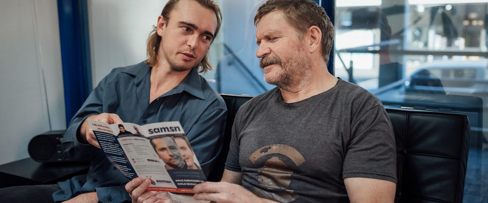 Two men sitting on a sofa reading a brochure and talking.